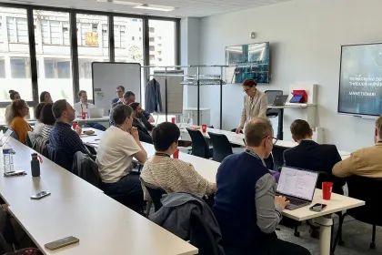 Conference room with people seated at tables while a presentation is shown on a screen.