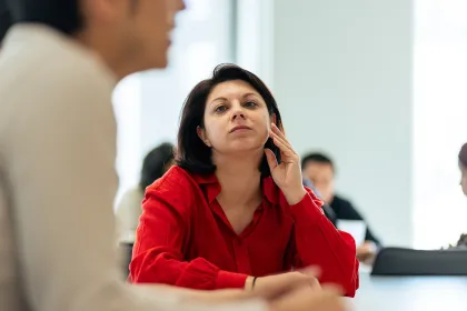 Une personne est assise à une table lors d’une réunion, regardant vers une autre personne au premier plan, avec plusieurs personnes et ordinateurs visibles en arrière-plan dans une salle lumineuse.