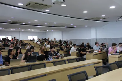 A large lecture hall with many people seated in rows, using laptops and tablets on wooden tables.