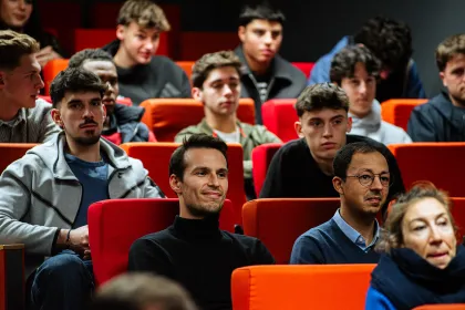 Public assis dans un amphithéâtre aux sièges rouges et orange lors d’une conférence ou d’un événement.