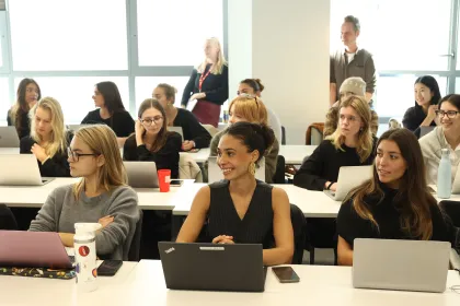 Several people are sitting around tables in a classroom, using laptops