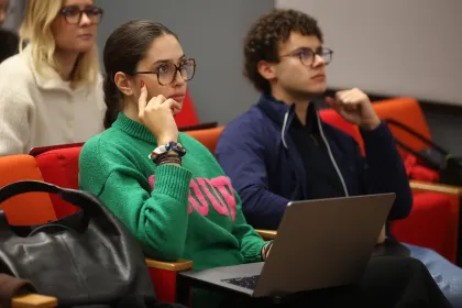 Several people are sitting around tables in a classroom, using laptops, while others are standing at the back of the room in front of large windows.