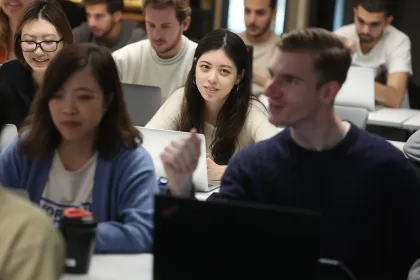 Plusieurs personnes sont assises dans une salle de cours, utilisant des ordinateurs portables autour de tables, avec des rangées de participants visibles à l’arrière-plan.