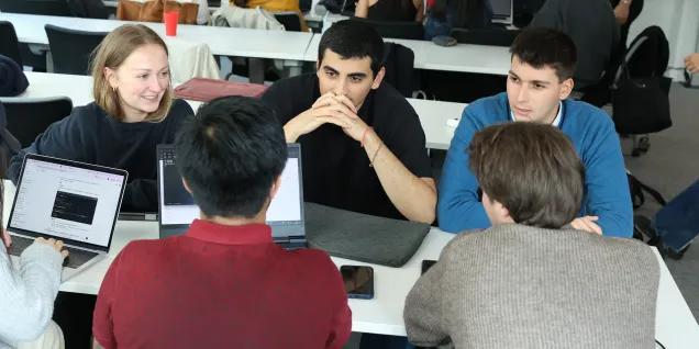 L'image montre un groupe de personnes rassemblées autour d'une table, travaillant sur des ordinateurs portables. Ils semblent être engagés dans une discussion ou une session de collaboration. Au fond, d'autres personnes et ordinateurs sont visibles, suggérant un environnement de travail ou d'apprentissage collaboratif. La scène dégage une atmosphère de convivialité et d'interaction.