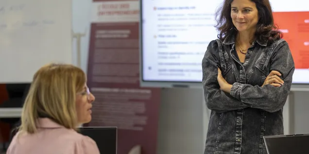 Une personne se tient debout avec les bras croisés, semblant expliquer ou présenter des informations, tandis qu'un autre participant, assis, l'écoute attentivement. En arrière-plan, un écran affiche des ressources ou des données pertinentes.