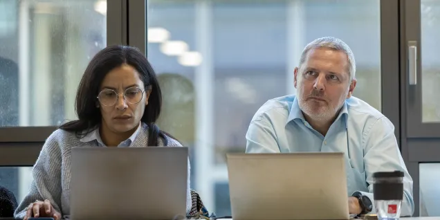 Deux personnes assises à une table de réunion, travaillant sur leurs ordinateurs portables. Un café et un verre d'eau sont présents sur la table, avec une grande fenêtre en arrière-plan laissant passer la lumière.