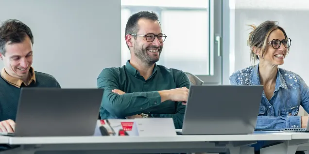 Groupe de trois personnes assises à une table, avec des ordinateurs portables, dans un cadre professionnel moderne. Ils semblent engagés dans une discussion ou une réunion.