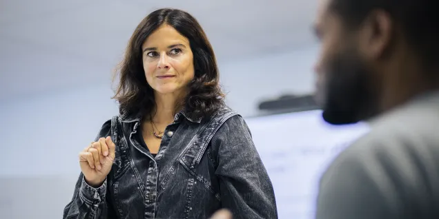 Une femme est debout, en train de parler, vêtue d'une chemise en jean. Elle semble engager une conversation ou présenter quelque chose, avec une main levée. En arrière-plan, on aperçoit un tableau ou un écran, suggérant un cadre de réunion ou de présentation. La scène dégage une atmosphère d'interaction professionnelle ou éducative.