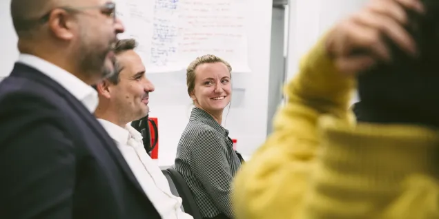 Dans cette image, on voit un groupe de personnes assises dans une salle, vraisemblablement lors d'une réunion ou d'une séance de brainstorming. On aperçoit des notes accrochées au mur en arrière-plan, ce qui indique qu'ils pourraient discuter de divers sujets. Les participants semblent attentifs et engagés dans la conversation. Les vêtements des personnes montrent une ambiance professionnelle, et l'environnement est moderne et minimaliste.