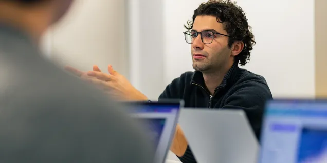 A meeting discussion with a participant gesturing, wearing a black sweater and glasses, in front of laptops in a modern work environment.