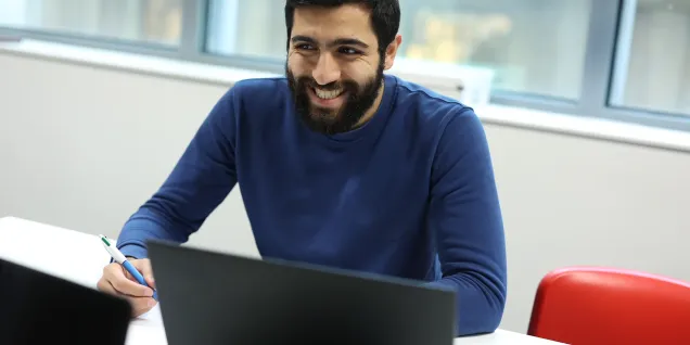 A man seated at a table, wearing a blue sweater. He is holding a pen and appears to be writing. On the table, there is a black laptop and another device. A window is visible in the background.
