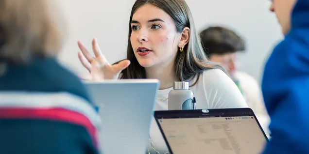 A group of students engaged in a discussion in a dynamic learning environment, with several open laptops on the table. One student is using gestures to express ideas while the others listen with attention. The atmosphere appears collaborative and active.