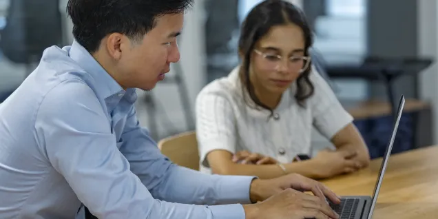 wo people seated at a wooden table working together on a laptop in a modern office setting. One person is typing on the keyboard while the other observes, creating a collaborative work atmosphere