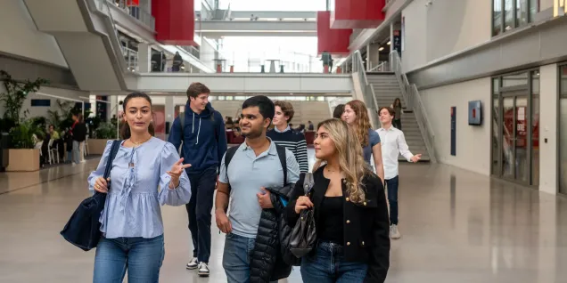 Un groupe d’étudiants et d’étudiantes d’emlyon marche dans le vaste atrium lumineux de l’école. L’espace présente de grandes baies vitrées, des passerelles, des escaliers et des éléments architecturaux rouges qui structurent le bâtiment. Le groupe avance au premier plan en discutant, tandis que d’autres personnes se déplacent à l’arrière-plan dans le cœur battant du campus.