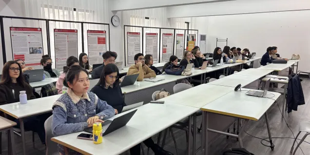 Un groupe d'étudiants attentifs dans un classroom moderne, assis à des tables en rangée avec des ordinateurs portables ouverts devant eux. Des affiches informatives se trouvent en arrière-plan, ajoutant un élément d'apprentissage visuel. L'atmosphère est studieuse et concentrée, mettant en avant l'engagement des étudiants dans leur éducation.