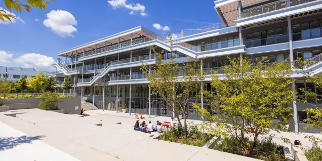 Vue du campus emlyon business school à Lyon, montrant un bâtiment moderne à plusieurs niveaux avec terrasses et passerelles extérieures, entouré d’arbres et d’espaces paysagers. Au premier plan, quelques personnes sont assises sur une esplanade en plein soleil.