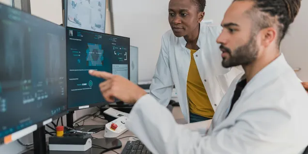 Two individuals in laboratory coats working at multiple monitors and reviewing scientific or medical data in a technical workspace