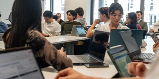 Salle de cours moderne avec étudiants travaillant sur ordinateurs portables lors d’une session académique, avec grandes fenêtres apportant de la lumière naturelle.
