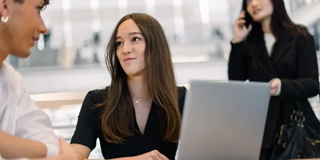 Three people in a modern workspace. A person in the foreground is wearing a black top and working on a laptop, while another person in the background is on the phone, and a third person stands next to them wearing a white shirt. The environment is bright and contemporary, with visible architectural elements.