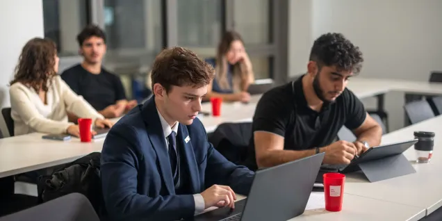 IMBA students at emlyon business school working on laptops during a class in a modern glass-walled classroom.