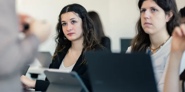 Personnes assises dans une salle de cours moderne, utilisant des ordinateurs portables et une tablette, attentives à une intervention en présentiel, dans un contexte d’enseignement supérieur et de formation.