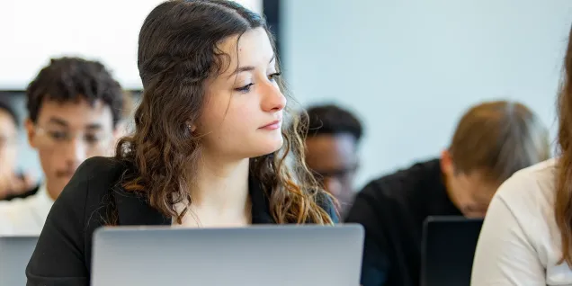 Student seated in a classroom using a laptop, with other students working in the background.