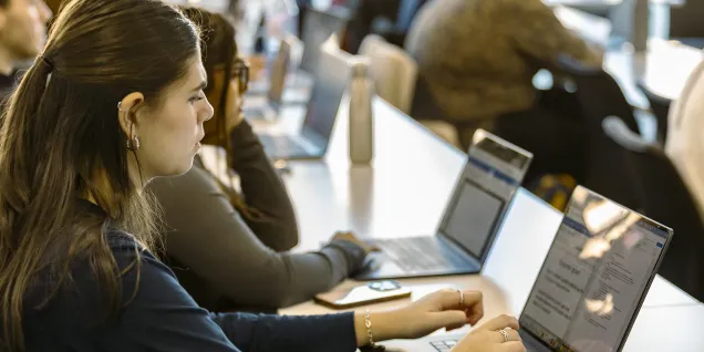 Students attending a class, seated at tables and working on laptops.