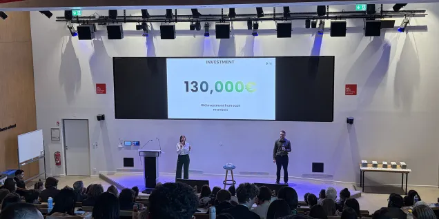 Wide view of a lecture hall during an event, with attendees seated in tiered rows and a presentation shown on a large screen.