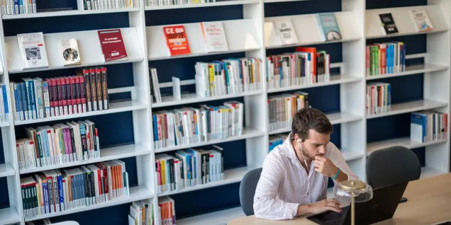 Une personne est assise à une table dans une bibliothèque, utilisant un ordinateur portable, avec des étagères de livres en arrière-plan et une lampe posée sur la table.