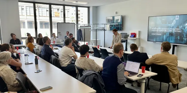 Conference room with people seated at tables while a presentation is shown on a screen.