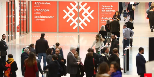 Personnes circulant dans le hall d’un campus universitaire, avec des panneaux institutionnels orange en arrière‑plan.