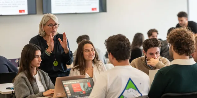 Classroom scene showing an instructor speaking with students gathered around laptops during a collaborative learning activity.