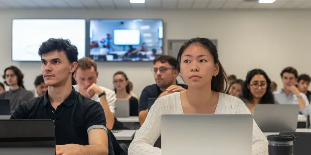 Students sitting in a classroom using laptops during a lecture, with a presentation displayed on screens in the background