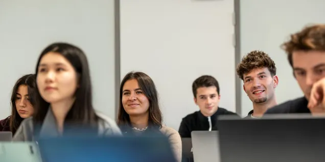 Un groupe de personnes assises dans une salle de classe utilise des ordinateurs portables, avec une porte fermée visible au fond de la pièce.