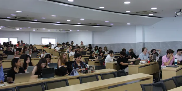 Une grande salle de cours en amphithéâtre avec de nombreuses personnes assises en rangées, utilisant des ordinateurs portables et des tablettes sur des tables en bois.