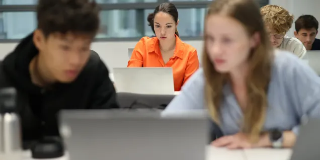 Students working on laptops in a classroom, with one person in focus in the center and others blurred in the foreground and background