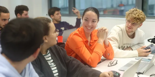 Étudiants collaborant autour d’ordinateurs portables en salle de classe, avec une personne en chemise orange au centre échangeant avec les autres