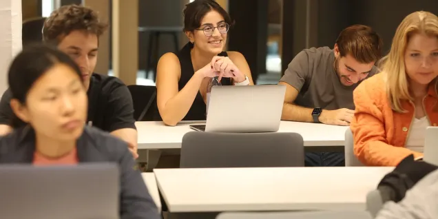 tudents using laptops in a classroom, with one person in focus at the center surrounded by classmates working at desks