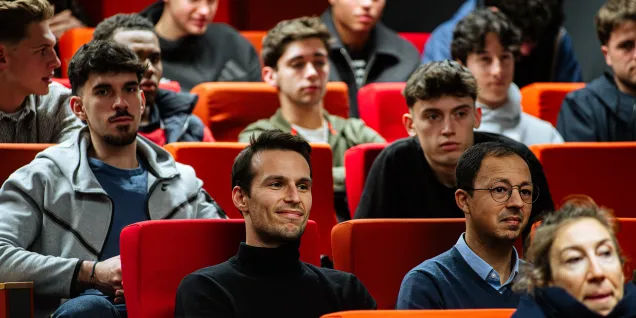 Public assis dans un amphithéâtre aux sièges rouges et orange lors d’une conférence ou d’un événement.