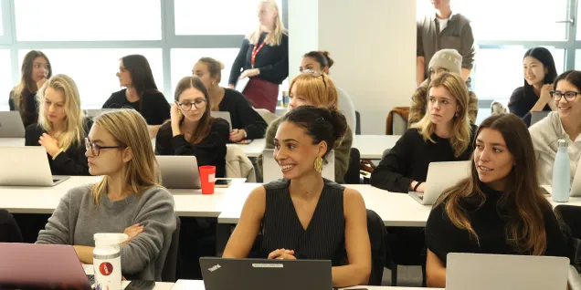 Several people are sitting around tables in a classroom, using laptops