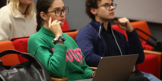 Plusieurs personnes sont assises dans une salle de cours autour de tables, utilisant des ordinateurs portables, tandis que d’autres se tiennent debout à l’arrière de la pièce devant de grandes fenêtres.