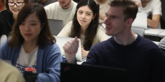 Several people are sitting in a classroom, using laptops at tables, with rows of participants visible in the background.