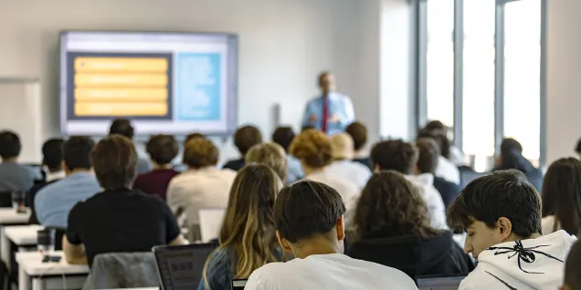 People are seated at tables in a lecture hall, some using laptops, while a presentation is projected onto a screen at the front of the room.