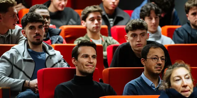 Participants assistant à une conférence ou à une présentation dans un amphithéâtre aux sièges rouges et orange.