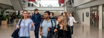 Un groupe d’étudiants et d’étudiantes d’emlyon marche dans le vaste atrium lumineux de l’école. L’espace présente de grandes baies vitrées, des passerelles, des escaliers et des éléments architecturaux rouges qui structurent le bâtiment. Le groupe avance au premier plan en discutant, tandis que d’autres personnes se déplacent à l’arrière-plan dans le cœur battant du campus.
