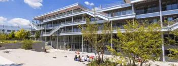 Vue du campus emlyon business school à Lyon, montrant un bâtiment moderne à plusieurs niveaux avec terrasses et passerelles extérieures, entouré d’arbres et d’espaces paysagers. Au premier plan, quelques personnes sont assises sur une esplanade en plein soleil.