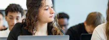 Étudiant·e assis·e dans une salle de classe, utilisant un ordinateur portable, avec d’autres étudiants travaillant en arrière-plan.
