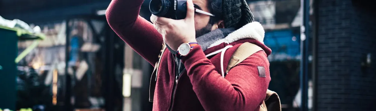 A person captures moments with a camera in an urban setting, dressed in a cozy red jacket and beanie.