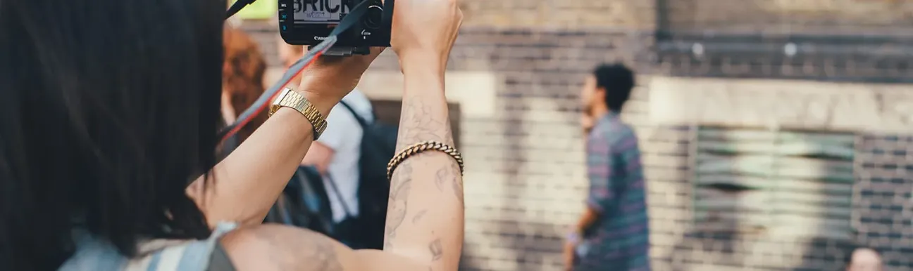 A photographer uses a digital SLR camera to capture an urban scene. People are seen moving in the background, and there is a brick environment.
