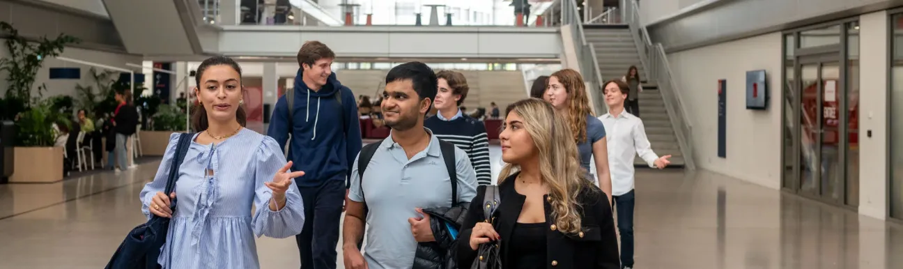 Un groupe d’étudiants et d’étudiantes d’emlyon marche dans le vaste atrium lumineux de l’école. L’espace présente de grandes baies vitrées, des passerelles, des escaliers et des éléments architecturaux rouges qui structurent le bâtiment. Le groupe avance au premier plan en discutant, tandis que d’autres personnes se déplacent à l’arrière-plan dans le cœur battant du campus.